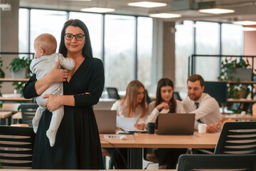 Little boy is in the hands of woman that is in black dress. Infant baby is in the office where group of people are working together