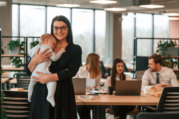 Little boy is in the hands of woman that is in black dress. Infant baby is in the office where group of people are working together