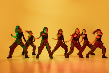 Group of little talented girls, children in sport style clothes dancing hip-hop against yellow studio background in neon light. Coordinated movements. Concept of childhood, hobby, sportive lifestyle