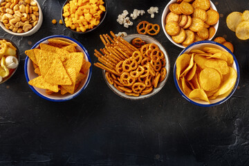 Salty snacks, party mix, overhead flat lay shot with copy space. An assortment of appetizers on a black background. Potato and tortilla chips, crackers, popcorn etc