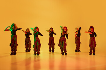 Group of little dancers, girls, children in stylish sportive clothes dancing against yellow studio background in neon light. Concept of childhood, hobby, sportive lifestyle, fashion, education