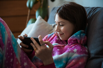 Obraz premium Happy teen girl looking at smartphone relaxing on couch at home, enjoying using online mobile apps technology, playing games on cell phone, checking messages or social media posts
