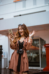 beautiful traveller with long brown hair waving her hand and looking at her phone while standing at front of the bulding