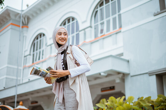 Female Traveller Looking Left While Standing At Front Of The Building Holding The Phone And The Maps