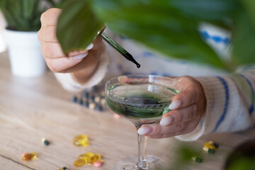a woman in a white sweater adds drops of chlorophyll to a glass of water standing next to palm trees