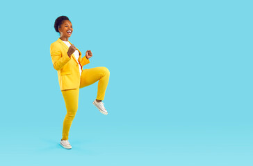 Fototapeta na wymiar Happy laughing woman dancing cheerfully showing yes gesture clenching her fists isolated on turquoise background, full length. Young stylish african american woman in yellow pants, jacket, sneakers.