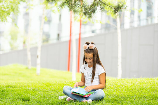 Cute little girl reading book on green grass near tree in park