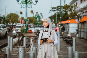 beautiful traveller with hijab holding the camera at her hand while standing at the sidewalk