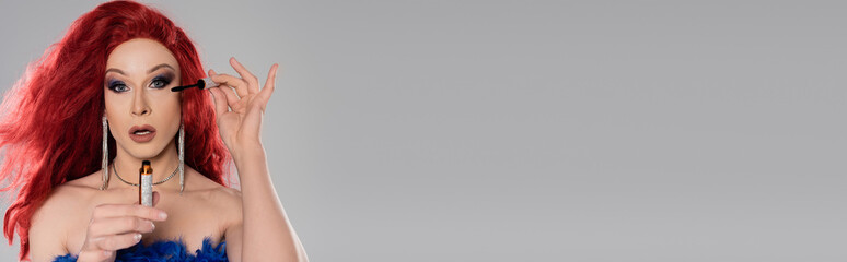 Drag queen in red wig holding mascara and looking at camera isolated on grey, banner.