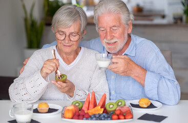 Smiling senior couple at home having breakfast together with muffin, milk and fresh seasonal fruit,...