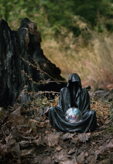 Magician mysterious black cloaked figure, Grim Reaper, with crystal ball in dark forest, blurred natural background. esoteric spiritual ritual for Samhain sabbat, Halloween holiday. Wiccan witchcraft
