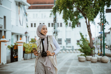 Fototapeta na wymiar female traveller with hijab smiling while walking through the sidewalk holding the phone and camera
