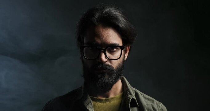 Closeup portrait of serious angry young man with beard and eyeglasses staring at camera against isolated black background