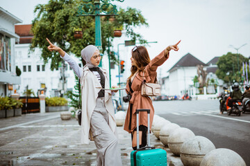 two female travellers pointing to the opposite way while standing together at the sidewalk bring their travel equipment
