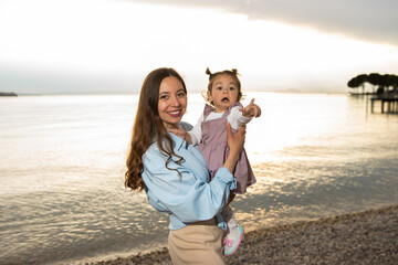 Young brunette mom in blue shirt holding her little toddler girl with two buns in velvet dress...