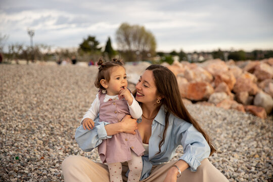 Young brunette mom in blue shirt sitting smiling with her little toddler girl with two buns in velvet dress at Italian Garda lake beach at sunset
