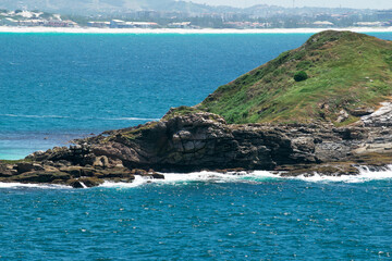 Island close to Praia dos Foguetes, close to the city of Cabo Frio, with many rocks and waves hitting them, blue sky, clear sea, mountains and part of the city in the background.