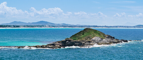 Island close to Praia dos Foguetes, close to the city of Cabo Frio, with many rocks and waves hitting them, clear sea, mountains and part of the city in the background.