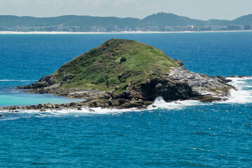 Island seen from Praia dos Foguetes, close to the city of Cabo Frio, with many rocks and waves hitting them, blue sky, clear sea and mountains in the background.