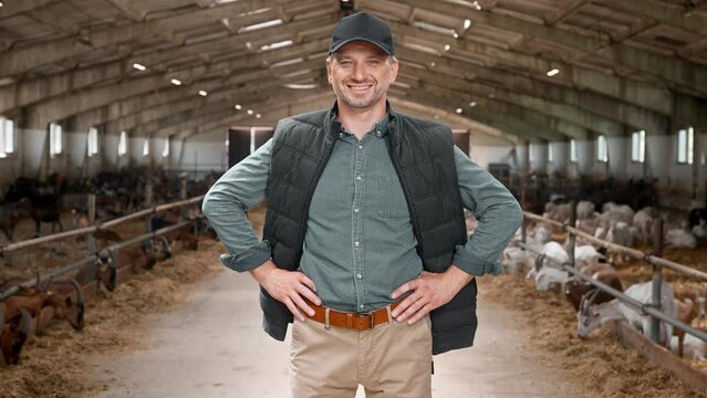 Attractive Caucasian farmer standing in barn. Positive man wearing cap and vest. Joyfully smiling and looking straight at camera. In background goats, sheep and lambs. Concept of agriculture business.