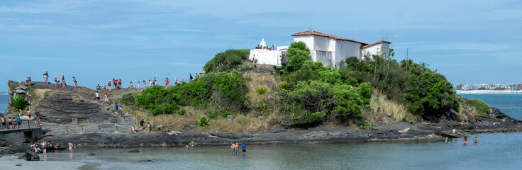 View of Praia do Forte, with the fort on top of the hill, blue sea, large rocks and cloudy sky.