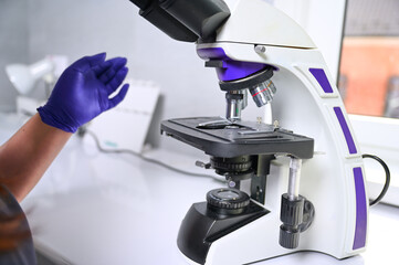 Close up view of laboratory female worker in blue gloves using microscope and examines fragments of...