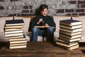 A studious young man dressed neatly, in round glasses is reading and studying among tall stacks of...