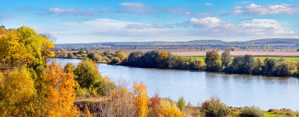 Autumn landscape with a forest on a hill near the river and a picturesque blue sky with white clouds in sunny weather
