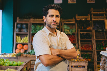 Middle-aged Latin man looking seriously at the camera with his arms crossed in a greengrocer's shop.