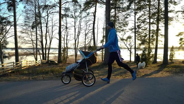 Male runner jogging with a pram baby carriage along an empty alley in a Park on the embankment on a evening in the setting sun rays in Finland