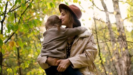 Hello September. A young mother in a brown hat and a little daughter walk in the city park in autumn. Mom holds the girl in her arms and hugs her tightly with love.