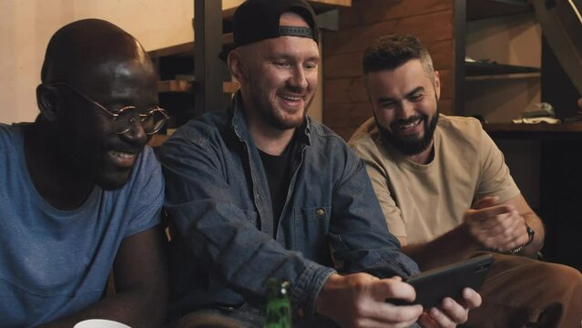 Medium shot of three multiracial male friends sitting on couch, drinking beer, man in middle showing video or photo to his friends, all laughing, Living room, stairs to first floor in backround