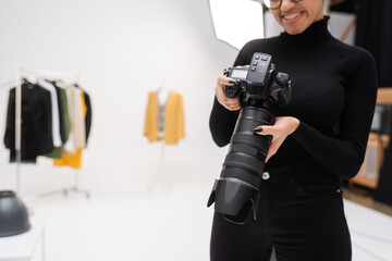 partial view of cheerful african american photographer in black turtleneck holding professional digital camera in blurred photo studio.
