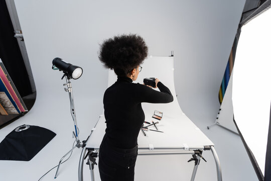 back view of african american photographer taking picture of beauty tools and decorative cosmetics on shooting table in photo studio.