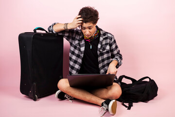 A lost and upset young traveller or tourist sitting on a baggage or spinner luggage is lost and looking at the laptop.