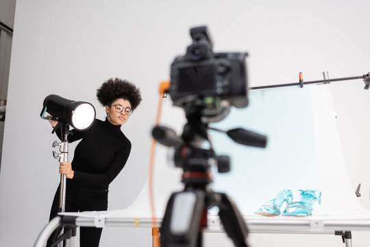 african american content manager adjusting strobe lamp near blurred digital camera and shooting table with stylish footwear in photo studio.