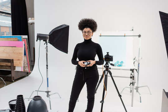 smiling african american content producer standing with exposure meter near digital camera and lighting equipment in modern photo studio.