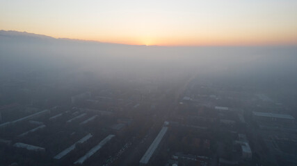 Epic gray smog is visible at sunset over the city. Aerial view from the drone of houses, roads, cars and parks. White clouds are illuminated by orange rays of the sun. The city is suffocating. Almaty