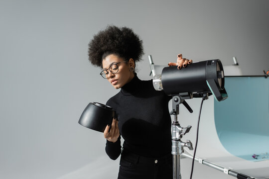 african american content maker in black turtleneck and eyeglasses holding detail of strobe lamp in photo studio.