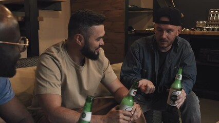 Medium cropped shot of three multiracial friends holding beer bottles, sitting on sofa, one speaking emotionally, counting on his fingers. Living room, sofa