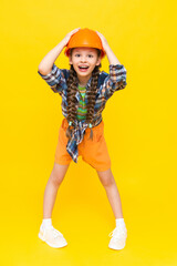 A little girl in an orange construction helmet is holding her head and smiling broadly, happily. The child is preparing for repairs in the nursery. In a mouthful. Vertical. Yellow isolated background