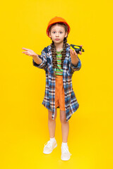 A little girl in an orange construction helmet holds a measuring tape. The concept of renovation in the children's room. The child is an engineer. Vertical. Full height. Yellow isolated background.