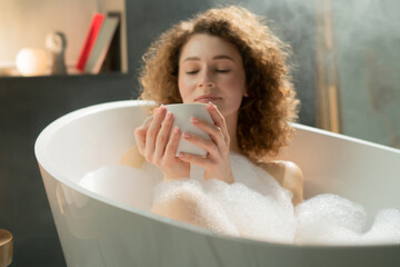 Young attractive woman enjoys hot fragrant tea while lying in a bath with foam and bubbles. A...