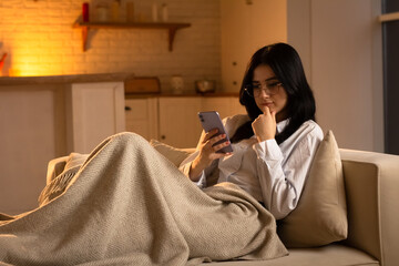 A young brunette uses a smartphone while lying on a sofa. The brunette lies on the couch and comes...