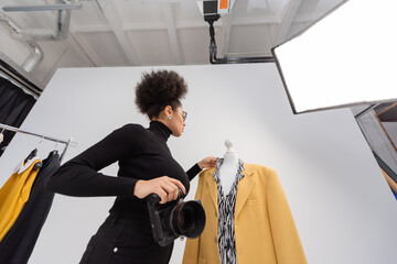 low angle view of african american photographer with digital camera adjusting stylish blazer in photo studio.