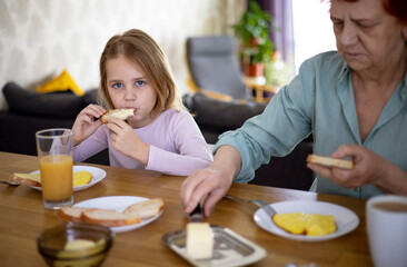 Obraz na płótnie Canvas A little blonde girl is having breakfast in the kitchen with her beloved grandmother. The child eats a healthy breakfast with an appetite. A woman smears bread with butter. Joy and good mood.