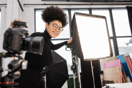 joyful african american content maker smiling at camera near floodlight and blurred digital camera in photo studio.