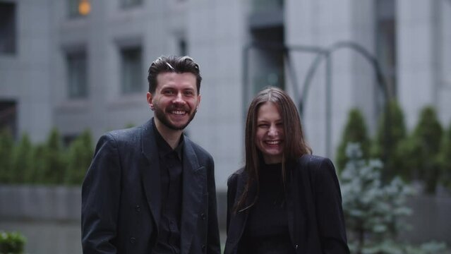 Two young men in strict black suits are laughing while looking at the camera. Business partners pose against the backdrop of a blurred building.
