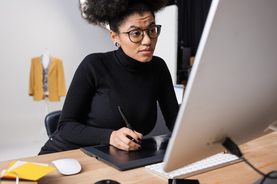 concentrated african american retoucher in eyeglasses and black turtleneck working on graphic tablet near computer monitor in photo studio. - Powered by Adobe