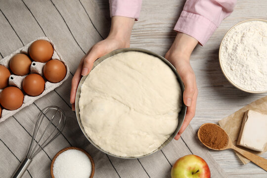 Woman holding bowl with fresh yeast dough and ingredients for cake on wooden table, closeup